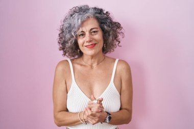 Pembe arka planda gri saçlı, elleri çapraz parmaklı, rahat ve neşeli gülümseyen orta yaşlı bir kadın. Başarı ve iyimserlik 