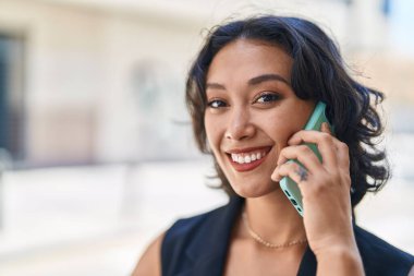 Genç, güzel, İspanyol bir kadın gülümsüyor ve caddede akıllı telefondan konuşuyor.