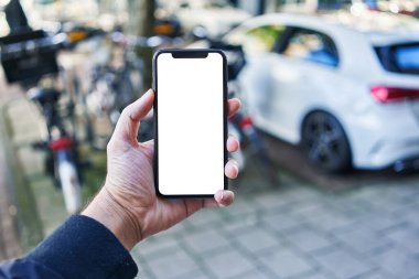 Akıllı telefon tutan adam araba parkında beyaz boş ekran gösteriyor.