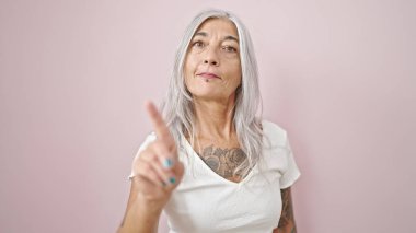 Orta yaşlı, gri saçlı kadın, izole edilmiş pembe arka planda parmağıyla hayır diyor.