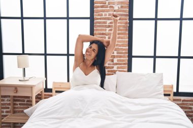 Middle age hispanic woman waking up stretching arms at bedroom