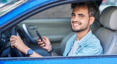 Young hispanic man using smartphone sitting on car at street