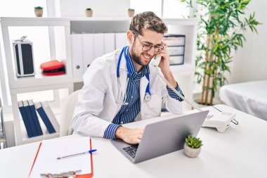 Genç doktor bilgisayarını kullanarak klinikte telefonla konuşuyor.