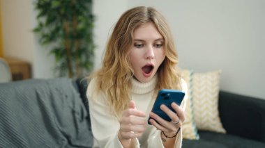 Young blonde woman using smartphone with surprise expression at home