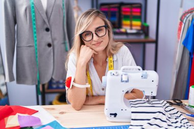 Young blonde woman tailor smiling confident sitting on table at tailor shop