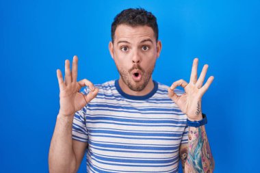 Mavi arka planda dikilen Latin asıllı genç adam şaşırmış ve şok olmuş görünüyor. Tamam sembolünü parmaklarıyla yapıyor. çılgın ifade 