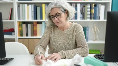 Middle age woman with grey hair teacher writing on notebook sitting on table at university classroom