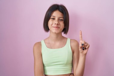 Pembe arka planda duran genç kız bir numarayı gösterip işaret ederken kendinden emin ve mutlu bir şekilde gülümsüyor.. 