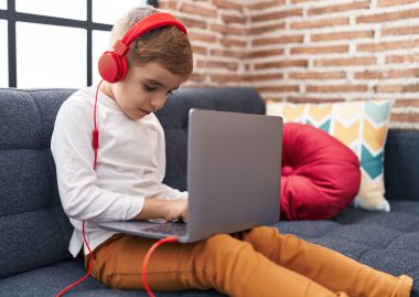 Adorable hispanic boy using laptop and headphones sitting on sofa at home