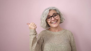 Middle age woman with grey hair smiling confident pointing to the side over isolated pink background