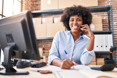 African american woman ecommerce business worker talking on smartphone writing on document at office