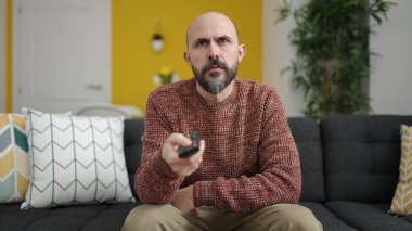 Genç kel adam televizyon seyrediyor. Evdeki sıkıcı ifadesiyle kanepede oturuyor.