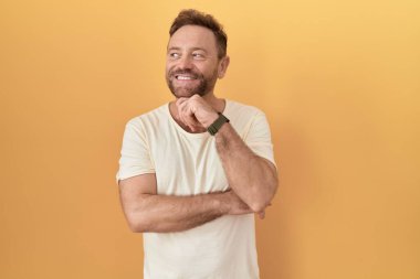 Sarı arka planda sakallı, eli çenesinde soru soran, düşünceli bir orta yaşlı adam. Gülümseyen ve düşünceli bir yüz. şüphe kavramı. 