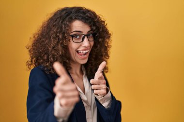 Sarı arka planda duran kıvırcık saçlı İspanyol kadın mutlu ve komik yüzlü parmaklarını kameraya doğrultuyor. iyi enerji ve titreşimler. 