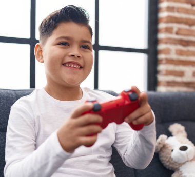 Adorable hispanic boy playing video game sitting on sofa at home