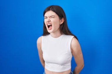 Young caucasian woman standing over blue background angry and mad screaming frustrated and furious, shouting with anger. rage and aggressive concept. 