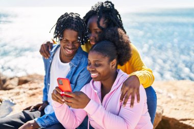 African american friends using smartphone sitting on rock at seaside