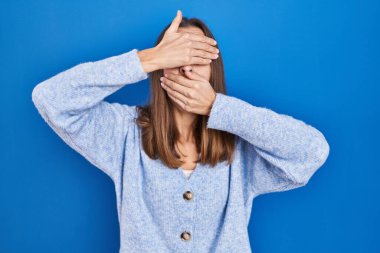 Mavi arka planda duran genç bir kadın gözlerini ve ağzını elleriyle kapatıyor, şaşırmış ve şok olmuş. duygu gizleme 