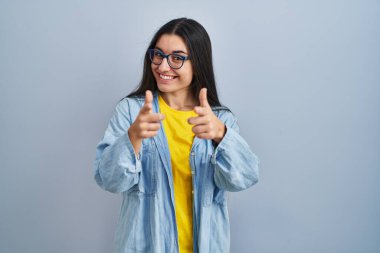 Mavi arka planda dikilen genç İspanyol kadın mutlu ve komik yüzlü parmaklarını kameraya doğrultuyor. iyi enerji ve titreşimler. 