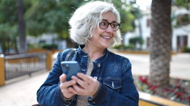 Middle age woman with grey hair using smartphone at park