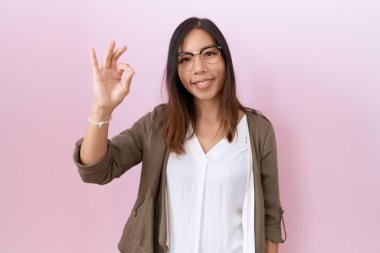 Orta yaşlı Çinli kadın pembe arka planda gözlük takıyor gülümsüyor ve el ve parmaklarıyla 