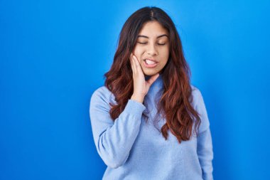 Mavi arka planda duran İspanyol genç bir kadın diş ağrısı ya da diş ağrısı yüzünden elleriyle dudaklarına dokunuyor. dişçi 