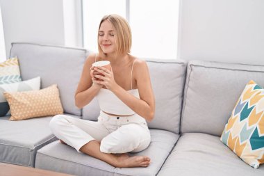 Young blonde woman drinking coffee sitting on sofa at home