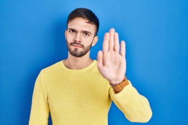 Hispanic man standing over blue background doing stop sing with palm of the hand. warning expression with negative and serious gesture on the face. 