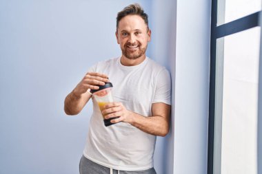 Genç beyaz adam, spor merkezinde protein içeceği içerek gülümsüyor.