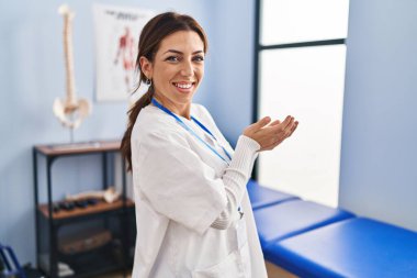 Ağrı kesici kliniğinde çalışan genç esmer kadın avuç içi açık bir şekilde fotokopi alanını gösteriyor, gülümseyerek mutlu bir gülümseme sunuyor. 