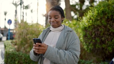 Parkta gülümseyen akıllı telefon kullanan Afrikalı Amerikalı kadın.