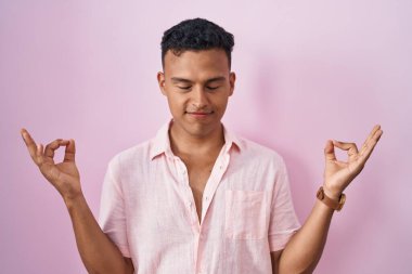 Pembe arka planda duran genç İspanyol adam rahatlamış ve gözleri kapalı gülümsüyor parmaklarıyla meditasyon hareketi yapıyor. yoga konsepti. 