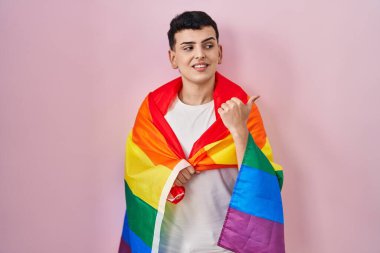 İkili olmayan kişi elinde gökkuşağı lgbtq bayrağı tutarken başparmağıyla yan tarafa bakıyor ve ağzı açık gülümsüyor. 
