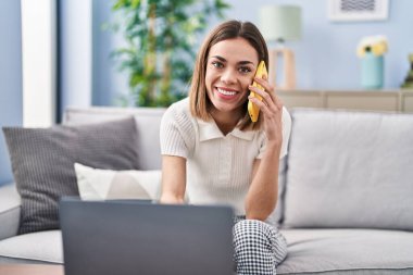 Genç, güzel, İspanyol bir kadın akıllı telefonuyla evde dizüstü bilgisayar kullanıyor.