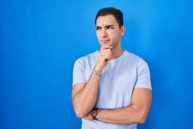 Young hispanic man standing over blue background thinking worried about a question, concerned and nervous with hand on chin  clipart