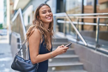 Genç, güzel, İspanyol bir kadın sokakta akıllı telefon kullanarak gülümsüyor.
