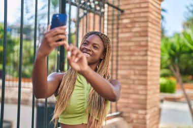 Afro-Amerikalı kadın gülümsüyor. Sokakta akıllı telefonun yanında özçekim yapıyor.