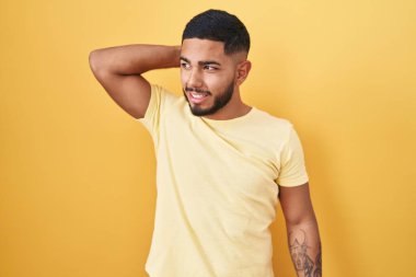 Young hispanic man standing over yellow background smiling confident touching hair with hand up gesture, posing attractive and fashionable 