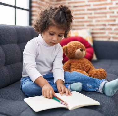 Adorable hispanic girl drawing on notebook sitting on sofa at home