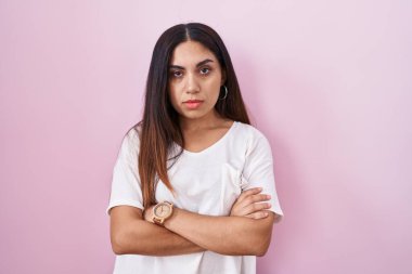 Pembe arka planda duran genç Arap kadın şüpheci ve gergin, yüzünde çapraz kollu tasvip etmeyen ifade. negatif kişi. 