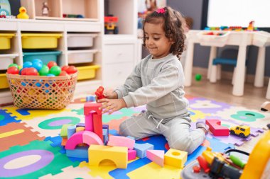 Adorable hispanic girl playing with construction blocks sitting on floor at kindergarten