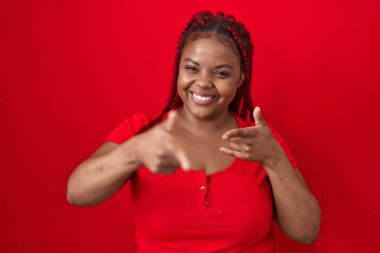 Kırmızı arka planda örülmüş saçları olan, mutlu ve komik yüzüyle kamerayı işaret eden Afro-Amerikalı bir kadın. iyi enerji ve titreşimler. 