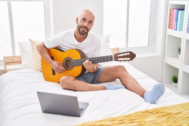 Genç kel bir adamın online klasik gitar dersi var. Yatak odasında oturuyor.