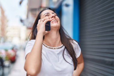 Genç, güzel, İspanyol bir kadın gülümsüyor ve caddede akıllı telefondan konuşuyor.