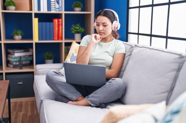 Evde dizüstü bilgisayar kullanan, kulaklık takan, ciddi yüzlü, eli çenesinde soru soran, kafası karışık, düşünceli genç bir kadın. 