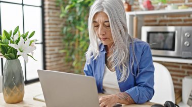 Orta yaşlı, gri saçlı bir kadın yemek odasındaki masada dizüstü bilgisayar kullanıyor.