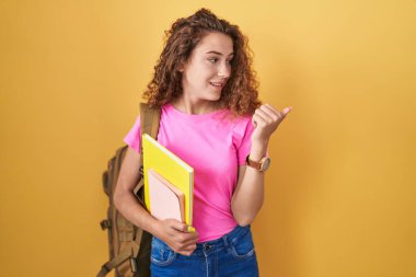 Sırt çantası takan, elinde kitaplarla gülümseyen, gülümseyen ve baş parmağıyla yüzünü gösteren beyaz bir kadın.. 