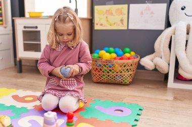 Anaokulunda yerde oturan sevimli sarışın kız oyuncaklarla oynuyor.