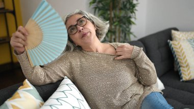 Middle age woman with grey hair sitting on sofa using handfan at home clipart