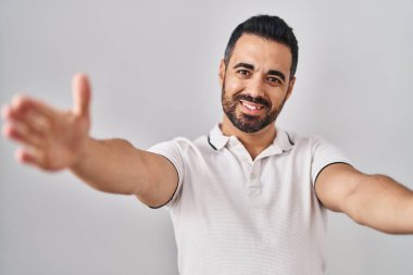 Beyaz arka plan üzerine günlük kıyafetler giyen sakallı genç İspanyol adam sarılmak için kollarını açmış kameraya bakıyor. Mutluluğu kucaklayan neşeli bir ifade.. 